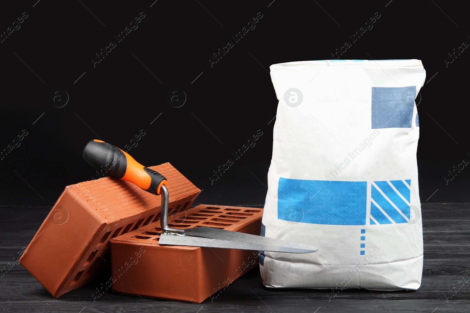 Photo of Red bricks, bag of cement powder and putty knife on dark wooden table. Building materials