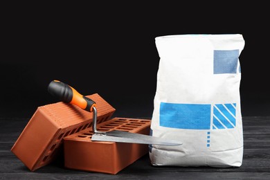 Red bricks, bag of cement powder and putty knife on dark wooden table. Building materials