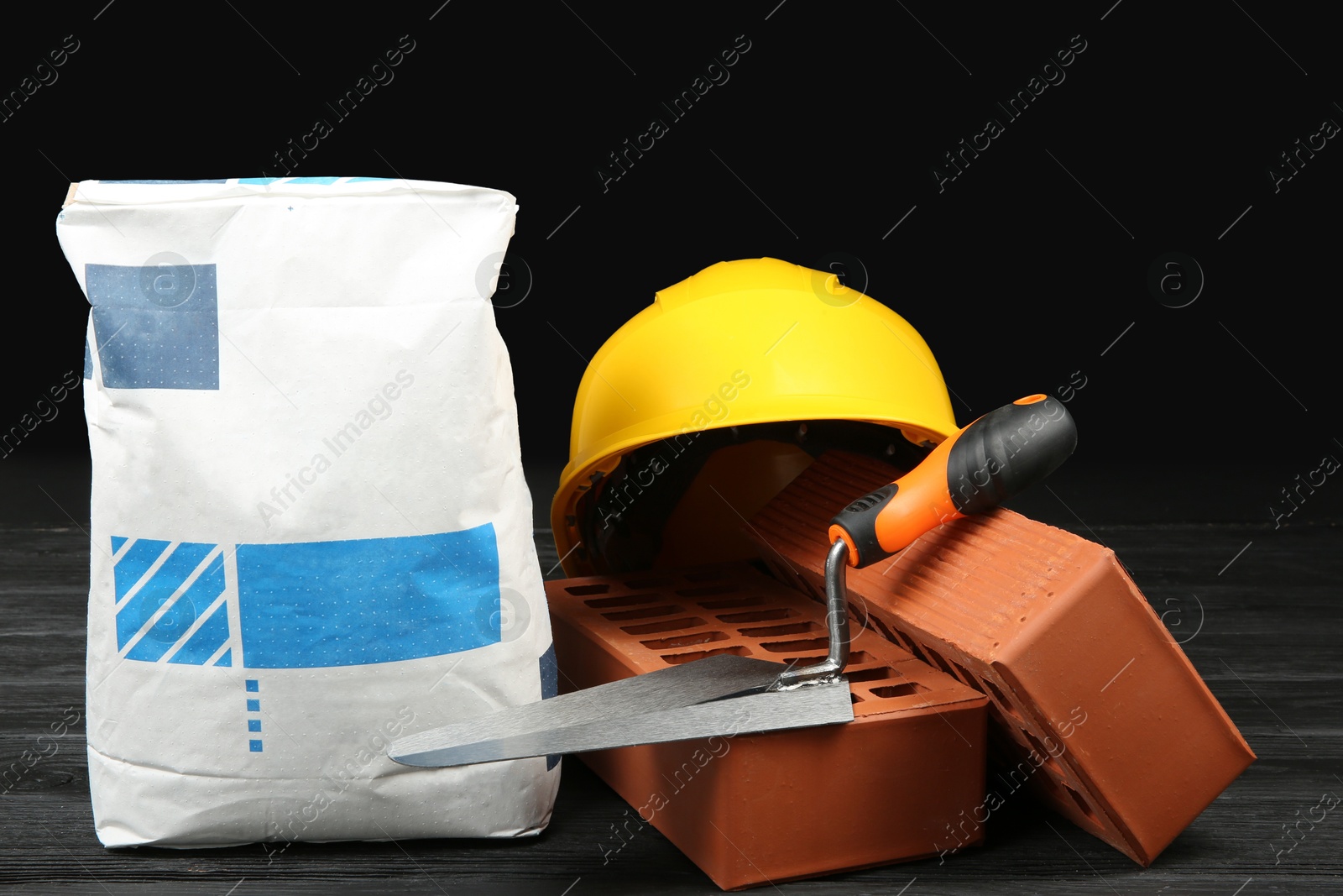 Photo of Red bricks, bag of cement powder, putty knife and yellow hard hat on dark wooden table. Building materials