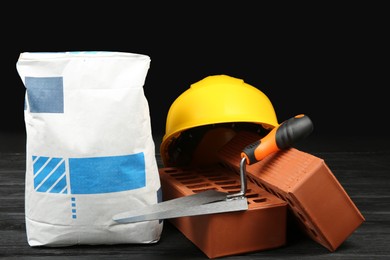 Photo of Red bricks, bag of cement powder, putty knife and yellow hard hat on dark wooden table. Building materials