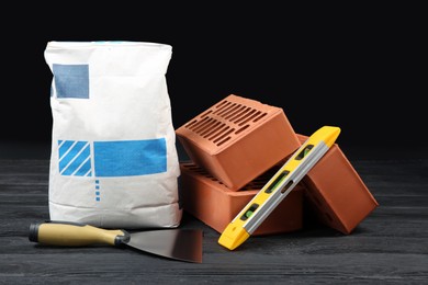 Photo of Red bricks, bag of cement powder, putty knife and spirit level on dark wooden table. Building materials and construction tools