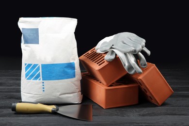 Photo of Red bricks, bag of cement powder, putty knife and rubber gloves on dark wooden table. Building materials