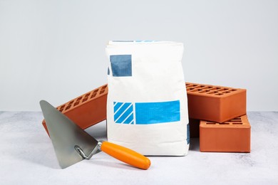 Photo of Red bricks, trowel and bag of cement on textured table against light background. Building material