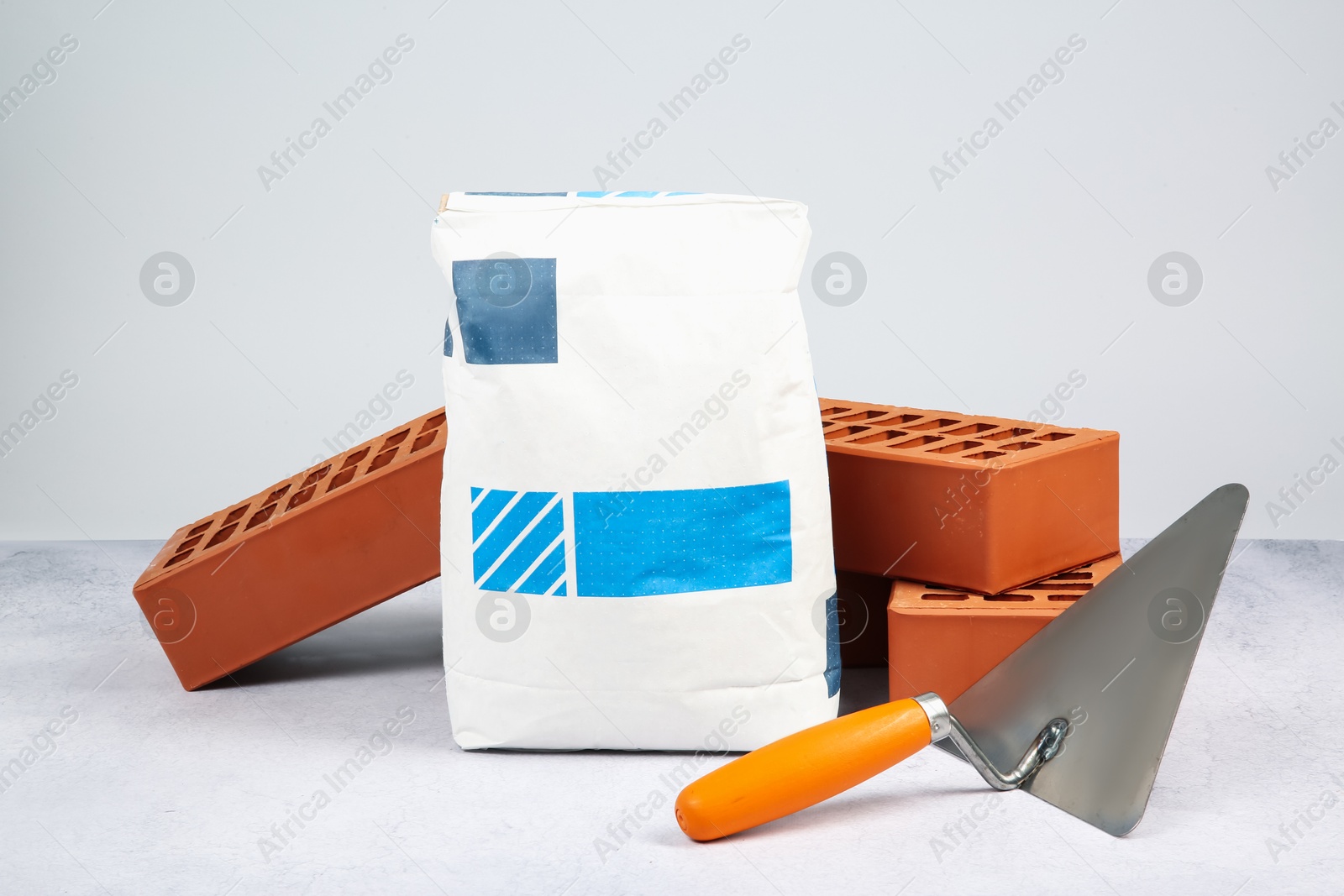 Photo of Red bricks, trowel and bag of cement on textured table against light background. Building material