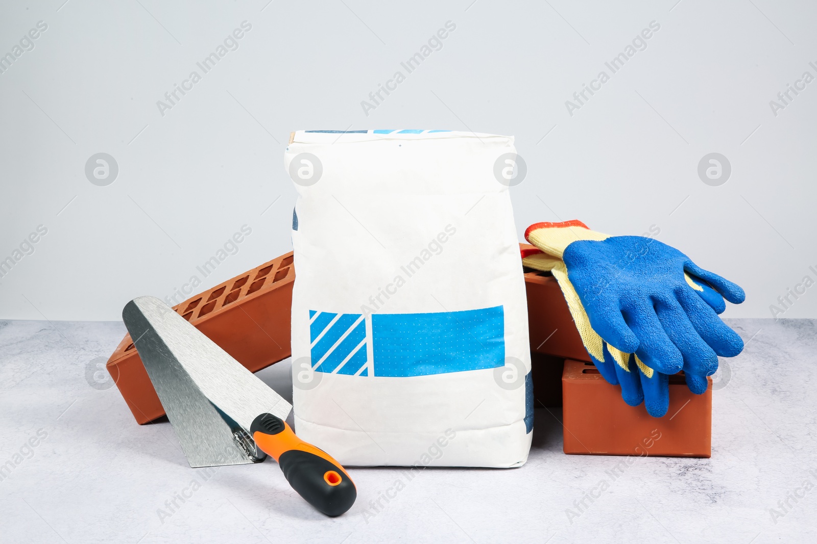 Photo of Red bricks, bag of cement, trowel and gloves on textured table against light background