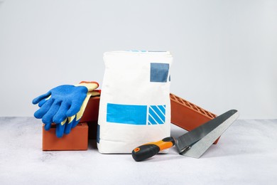 Photo of Red bricks, bag of cement, trowel and gloves on textured table against light background