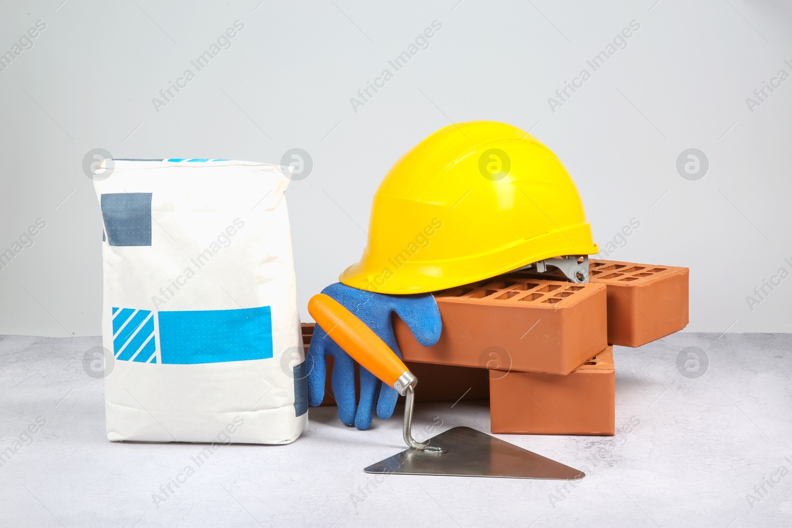 Photo of Red bricks, bag of cement, trowel and glove on textured table against light background