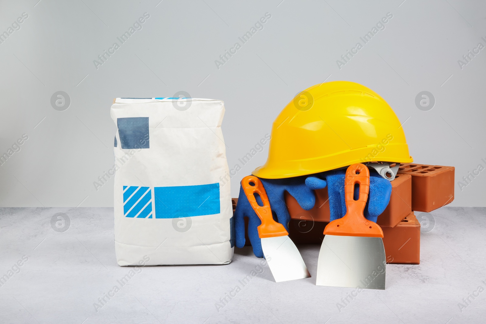 Photo of Red bricks, bag of cement, trowels, hard hat and gloves on textured table against light background