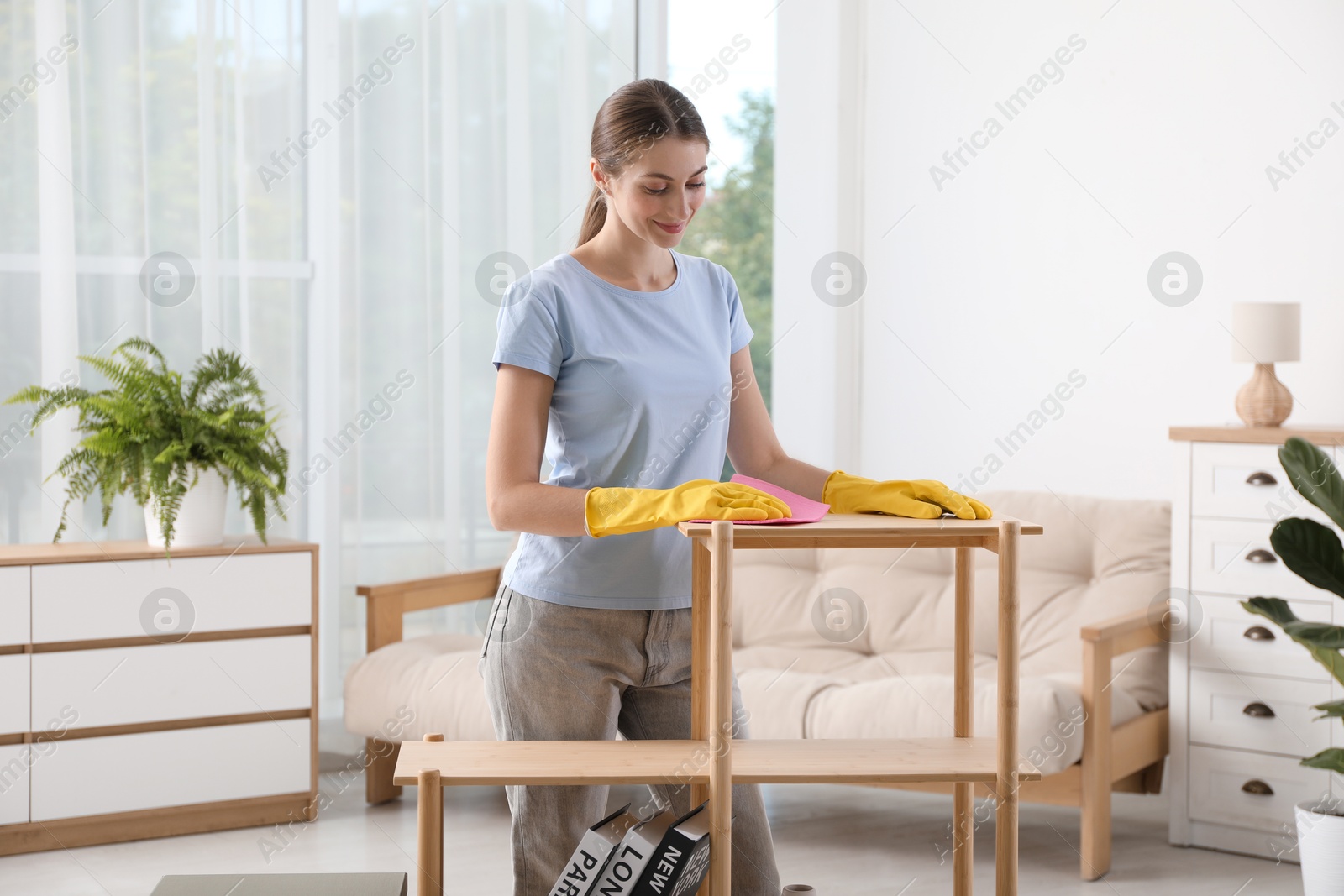 Photo of Young woman cleaning wooden shelf with rag at home