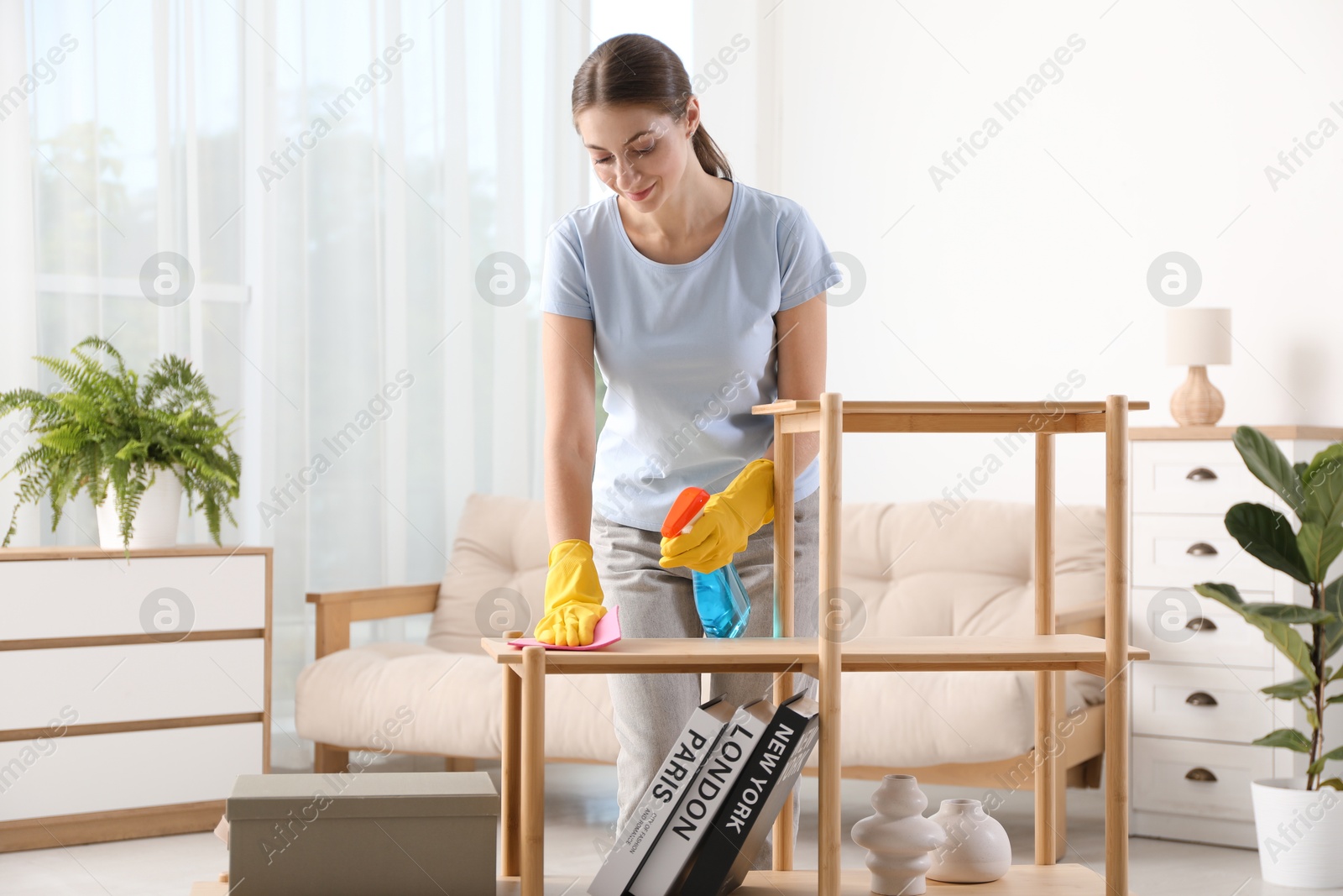 Photo of Young woman cleaning wooden shelf with rag and spray at home