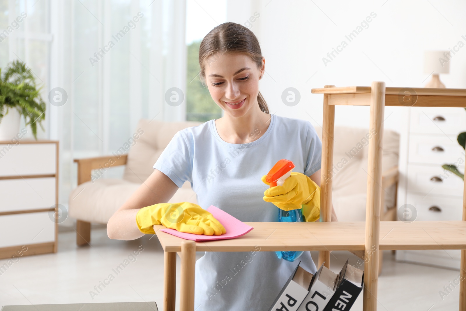 Photo of Young woman cleaning wooden shelf with rag and spray at home