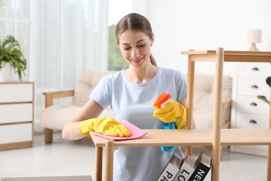 Young woman cleaning wooden shelf with rag and spray at home