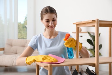 Photo of Young woman cleaning wooden shelf with rag and spray at home