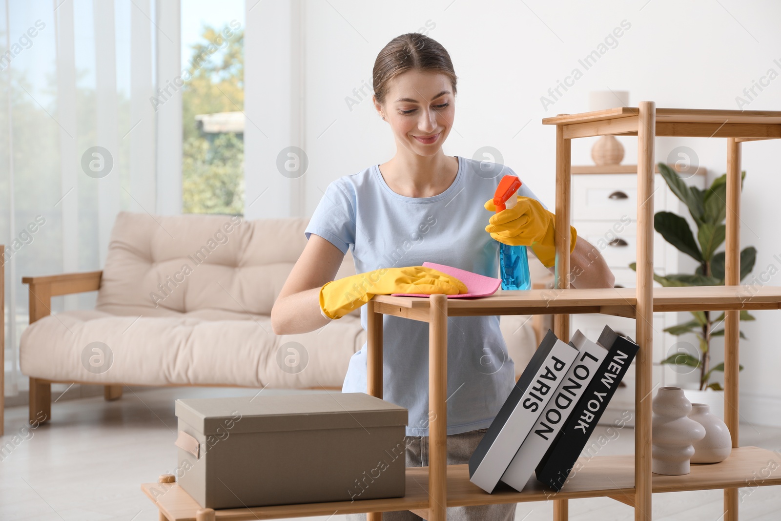 Photo of Young woman cleaning wooden shelf with rag and spray at home