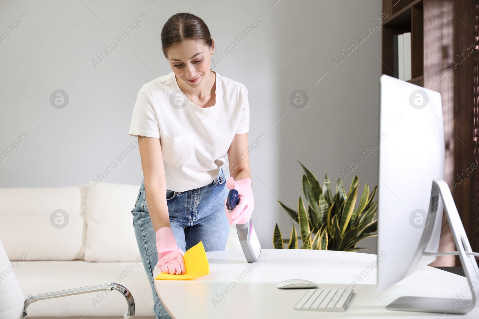 Photo of Young woman cleaning table with rag and spray in office