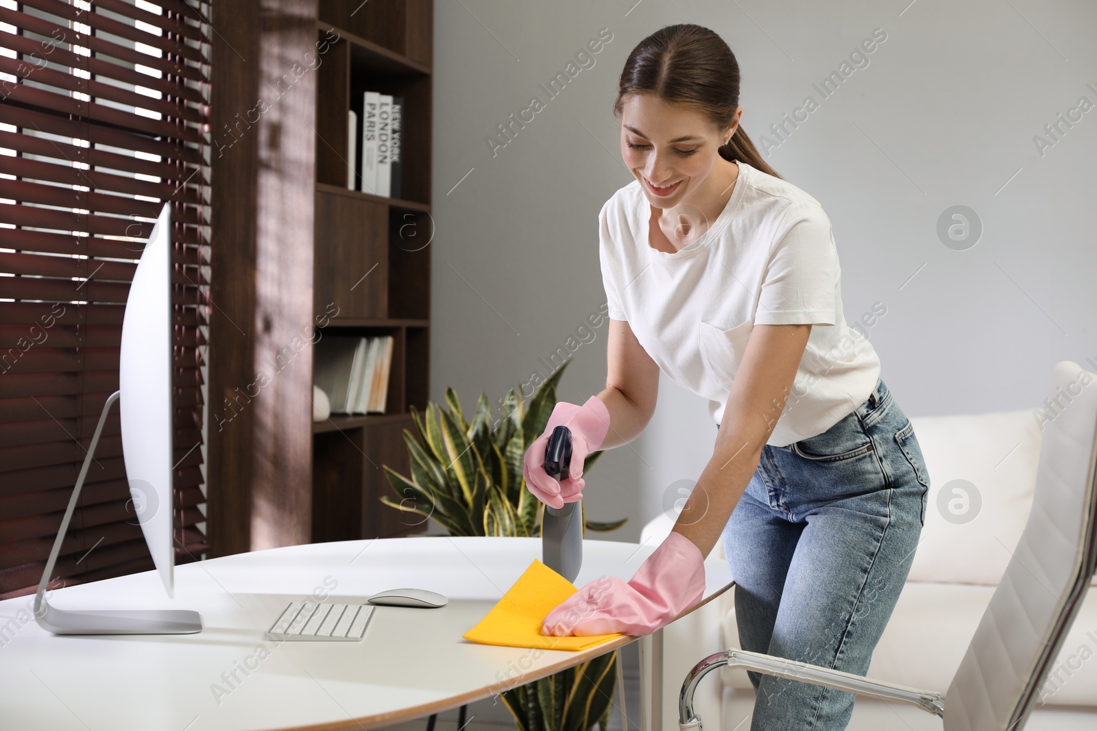 Photo of Young woman cleaning table with rag and spray in office