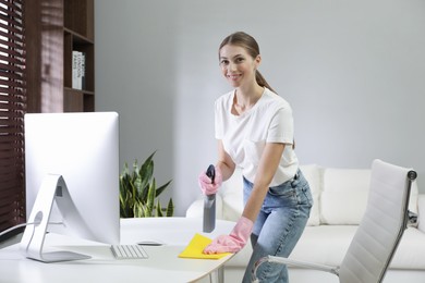 Photo of Young woman cleaning table with rag and spray in office