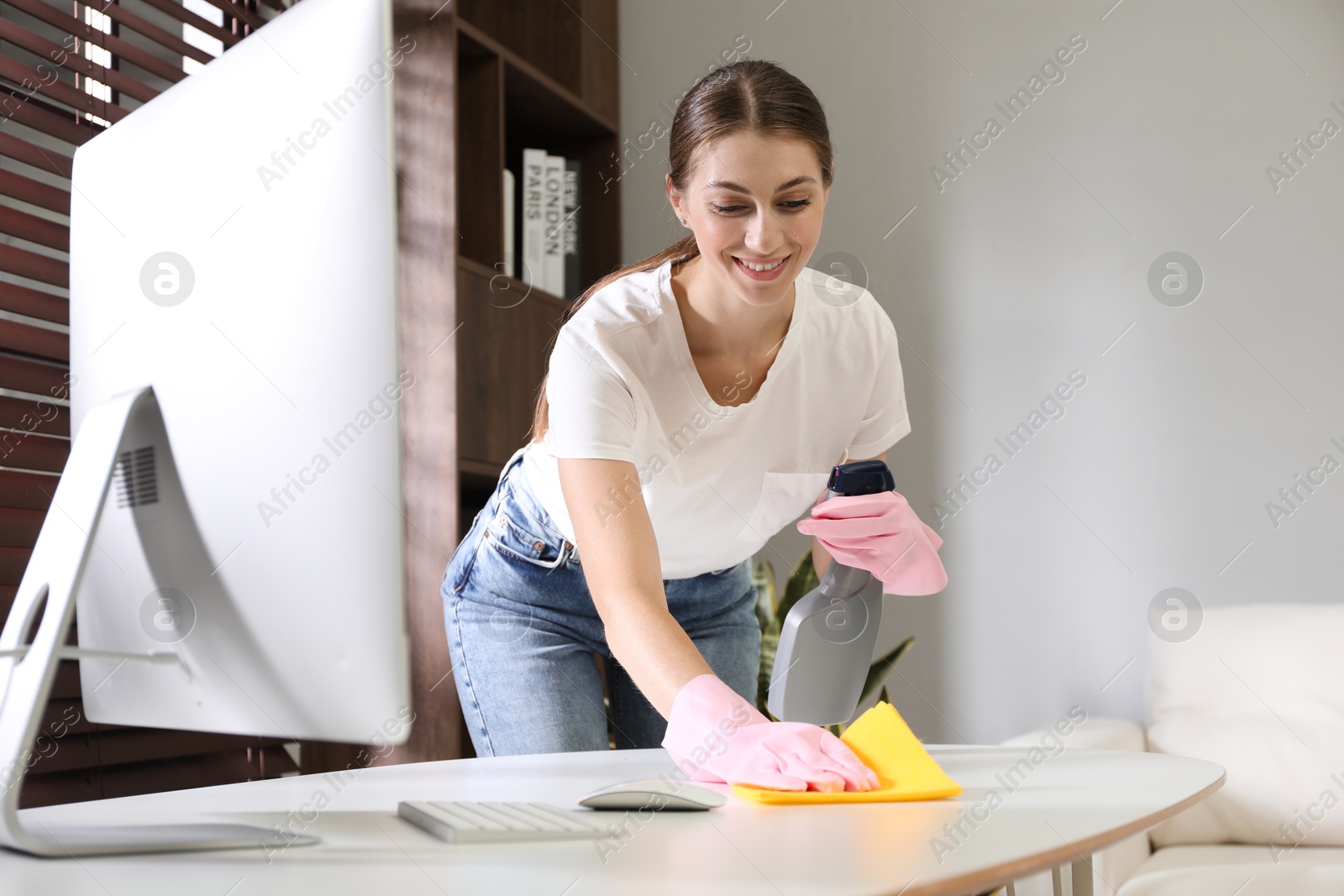 Photo of Young woman cleaning table with rag and spray in office