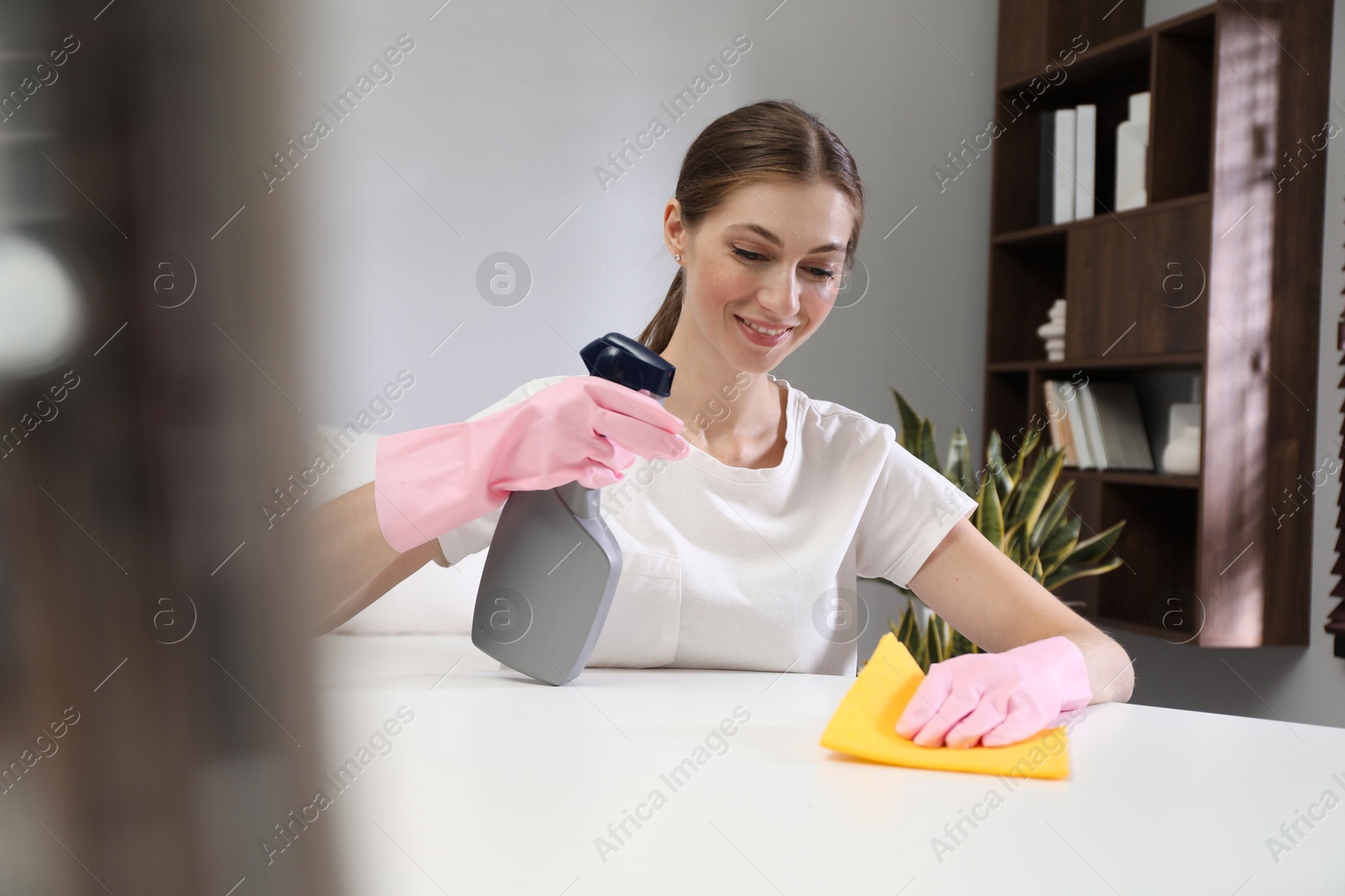 Photo of Young woman cleaning table with rag and spray in office