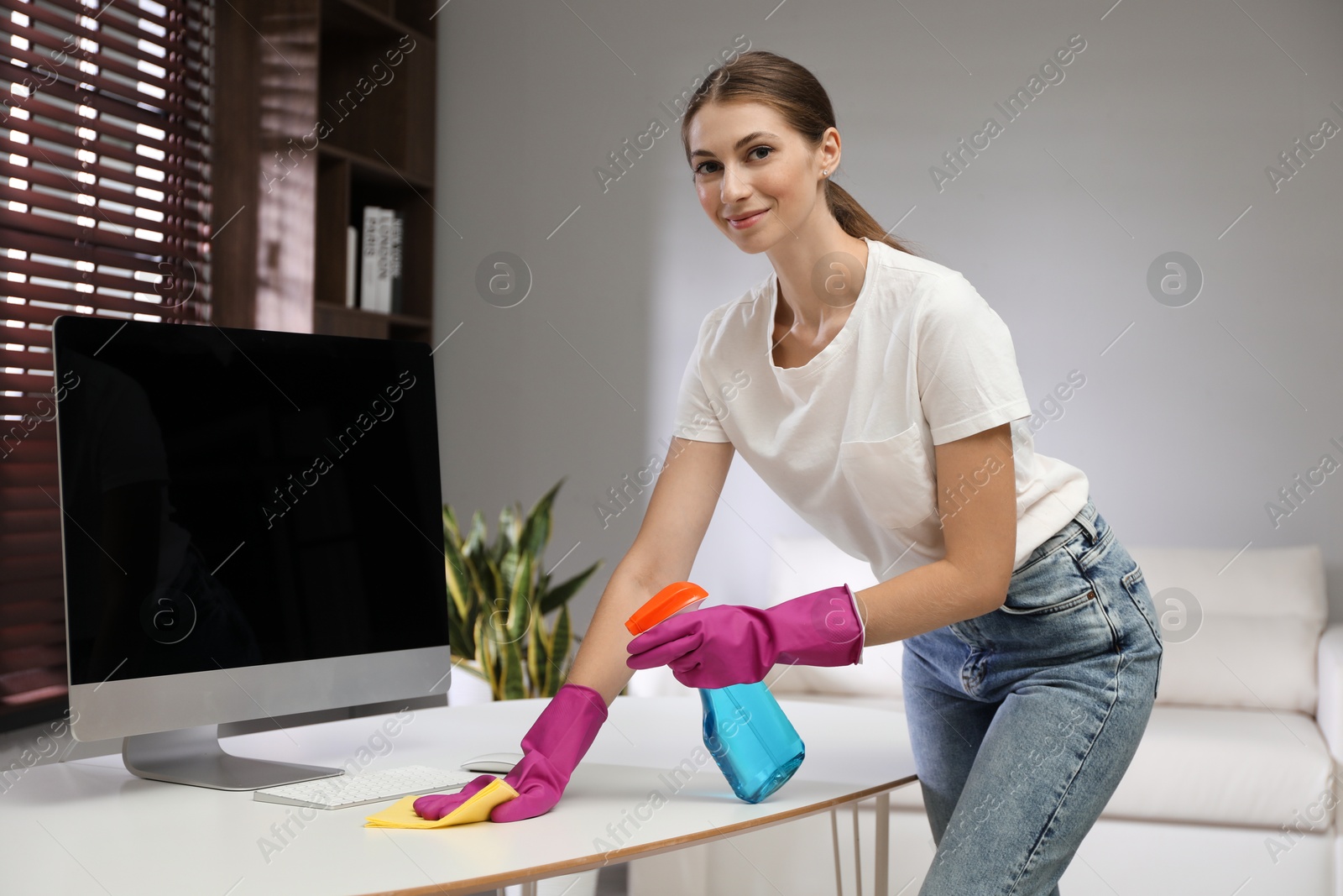 Photo of Young woman cleaning table with rag and spray in office