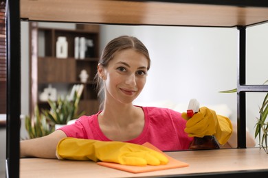 Young woman cleaning wooden shelf with rag and spray in office