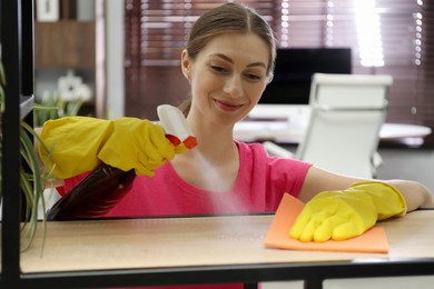 Young woman cleaning wooden shelf with rag and spray in office