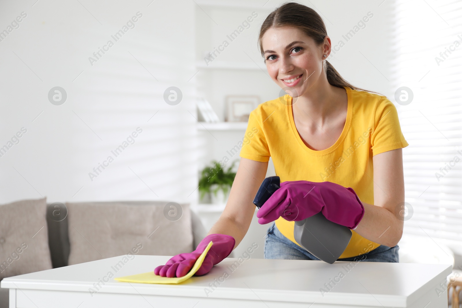 Photo of Young woman cleaning table with rag and spray in office