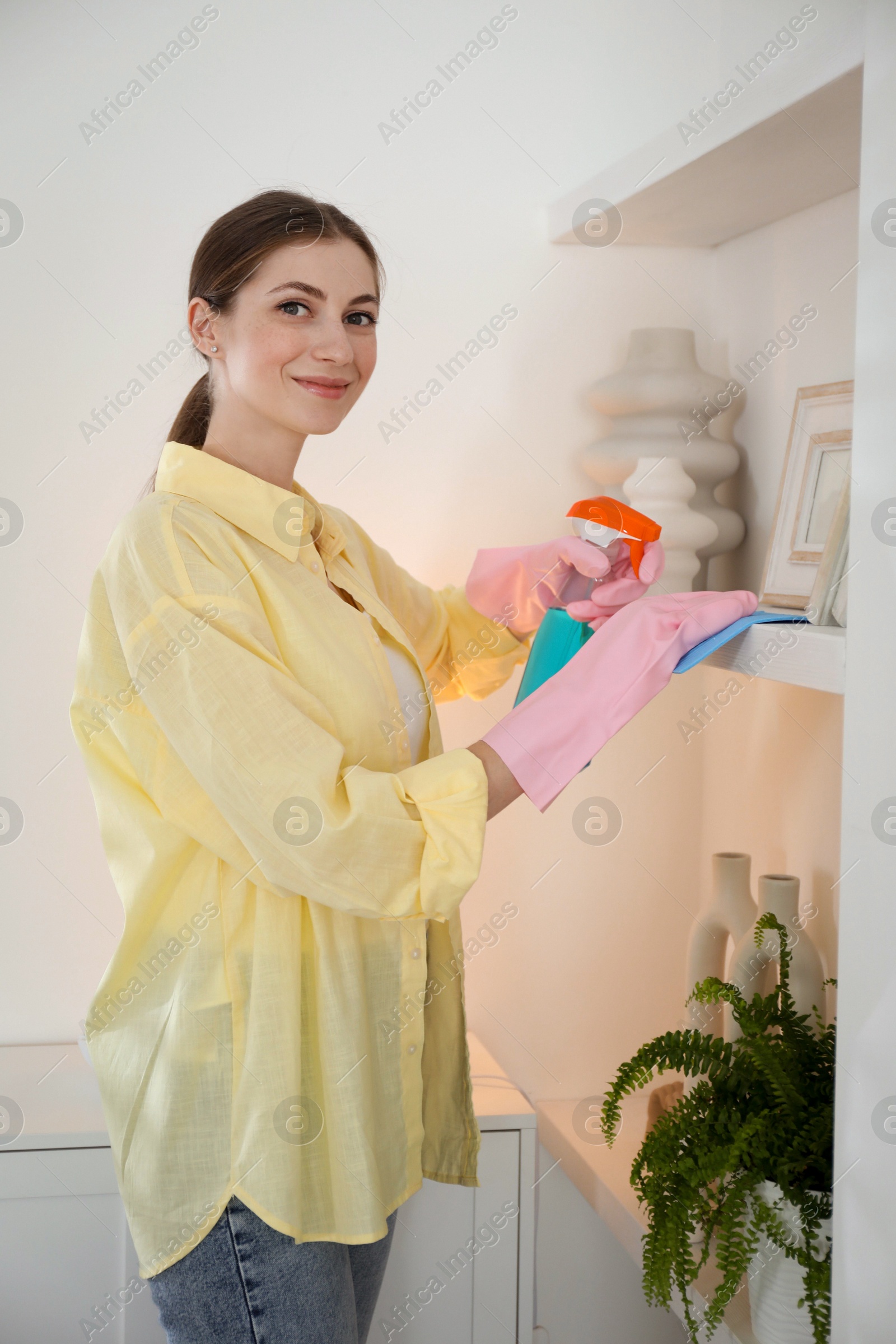 Photo of Young woman cleaning shelf with rag and spray at home