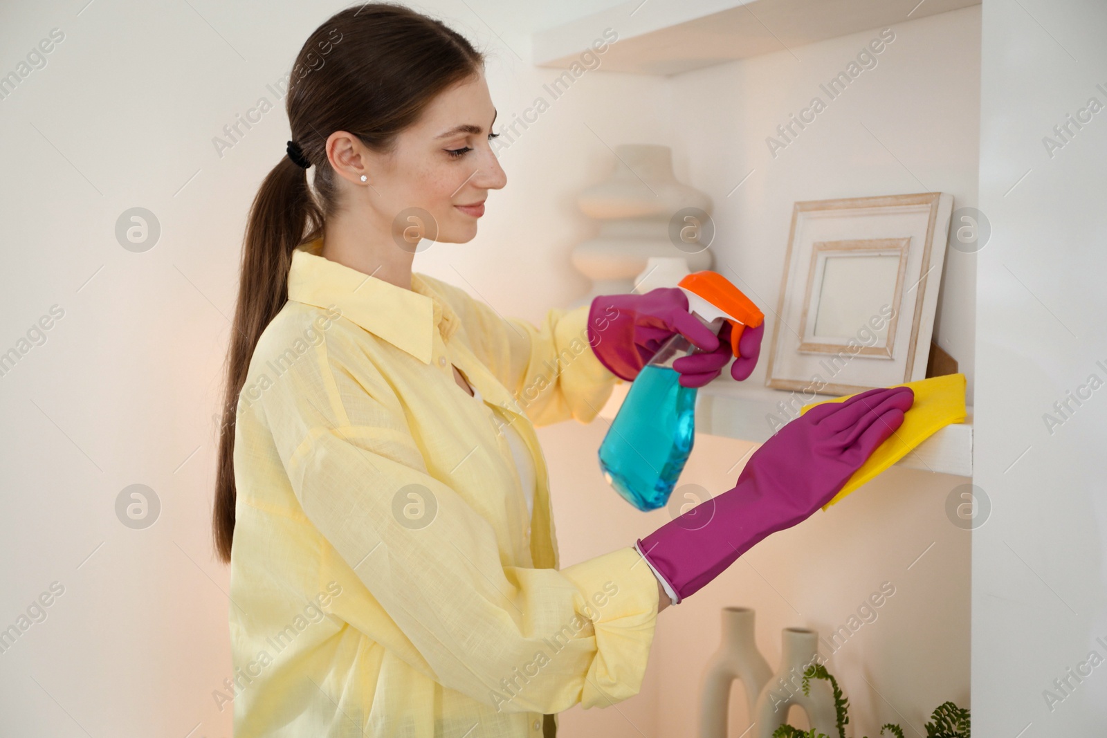 Photo of Young woman cleaning shelf with rag and spray at home