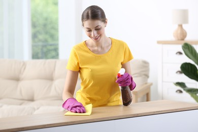 Young woman cleaning wooden table with rag and spray at home