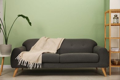 Stylish sofa with blanket, shelving unit and houseplant near green wall indoors