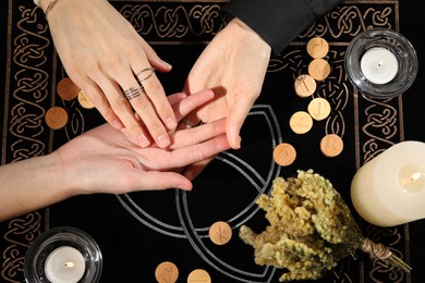 Photo of Fortuneteller reading woman's hand at table, top view. Wooden runes and burning candles on divination mat