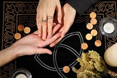 Photo of Fortuneteller reading woman's hand at table, top view. Wooden runes and burning candles on divination mat