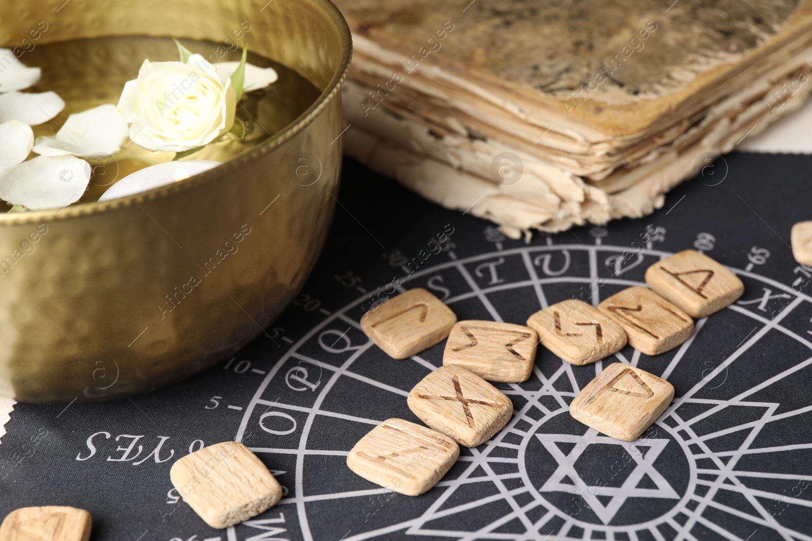 Photo of Wooden runes, old book and bowl with flower petals in water on divination mat, closeup