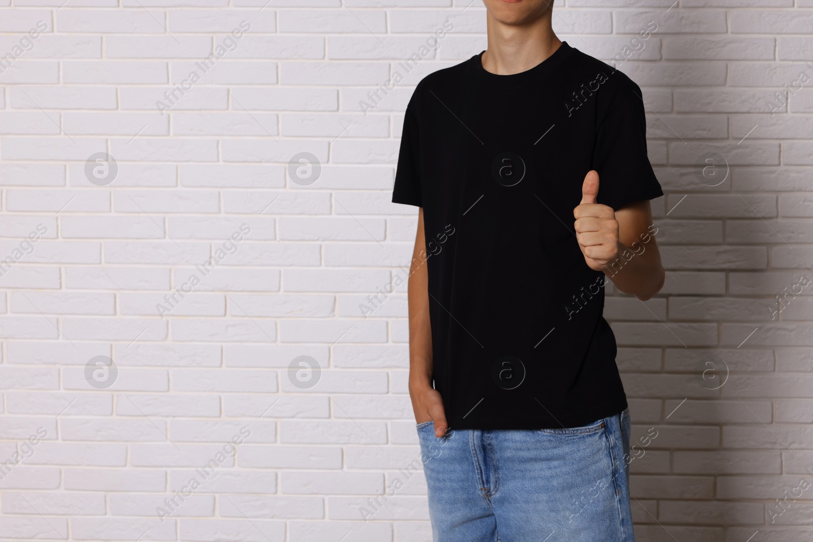 Photo of Teenage boy wearing black t-shirt and showing thumbs up near white brick wall, closeup. Space for text