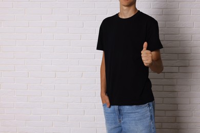 Photo of Teenage boy wearing black t-shirt and showing thumbs up near white brick wall, closeup. Space for text