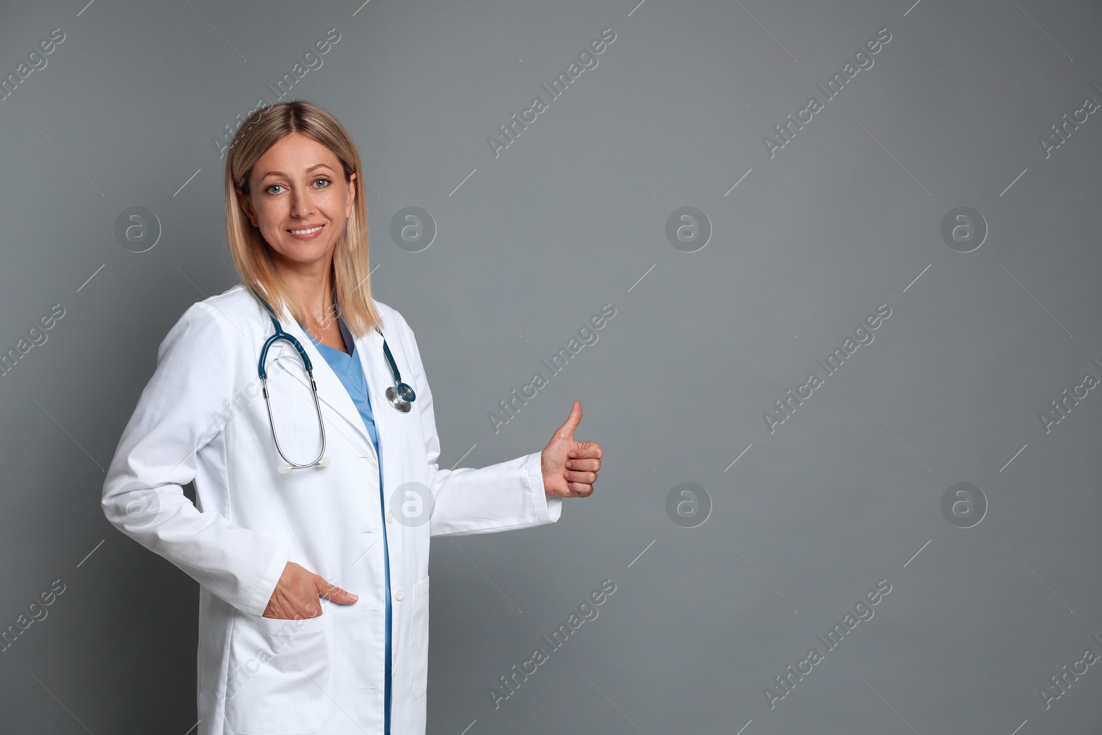 Photo of Doctor in medical coat with stethoscope showing thumbs up on grey background, space for text