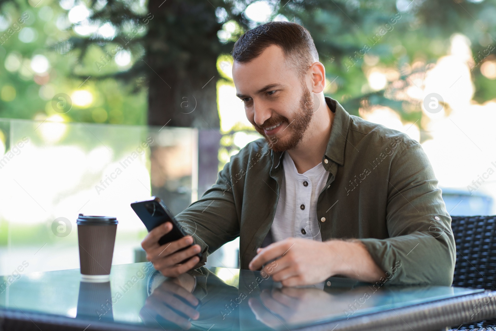 Photo of Happy man with paper cup using smartphone at outdoor cafe