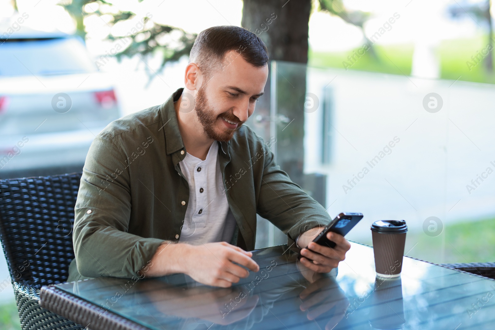 Photo of Happy man with paper cup using smartphone at outdoor cafe