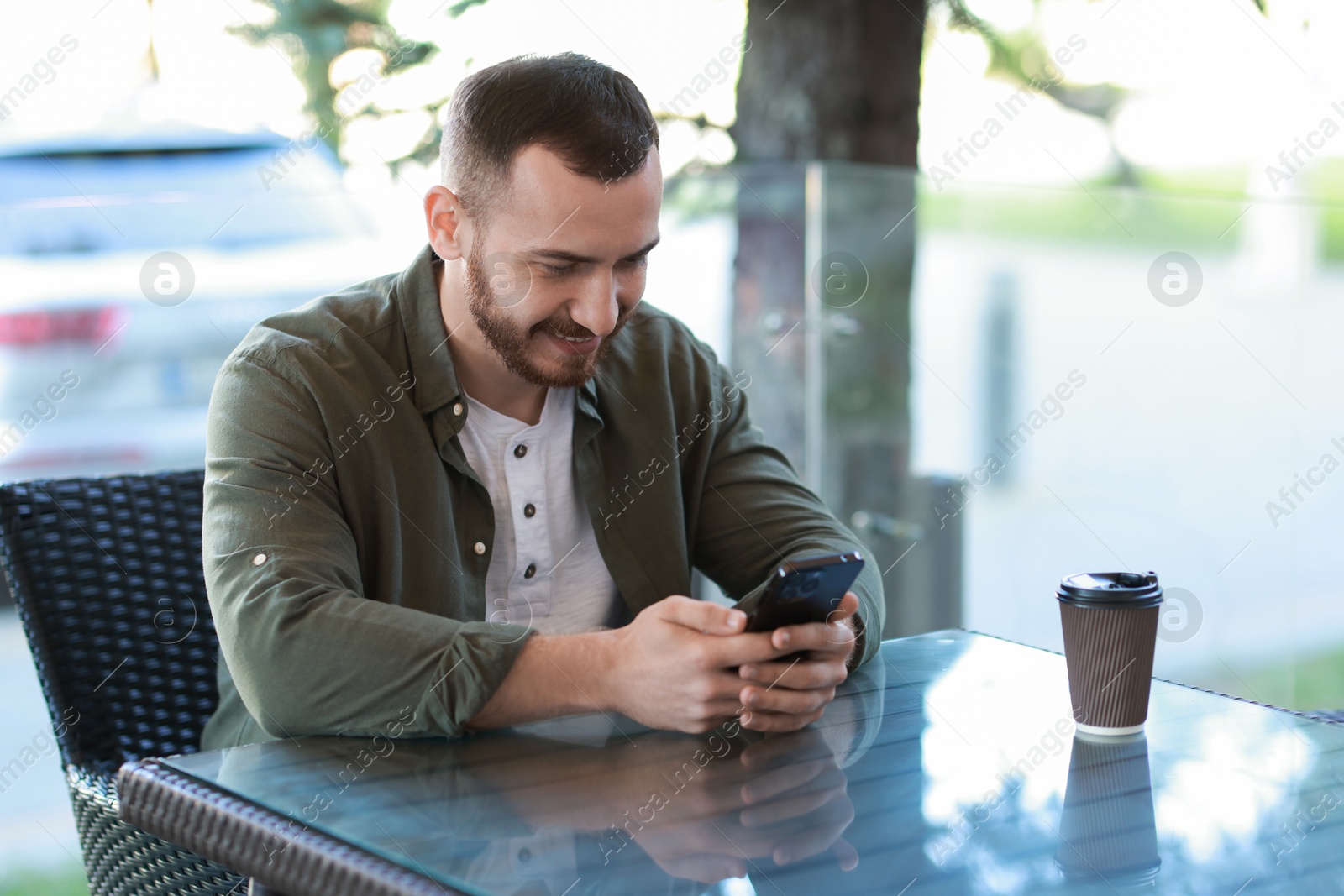 Photo of Happy man with paper cup using smartphone at outdoor cafe