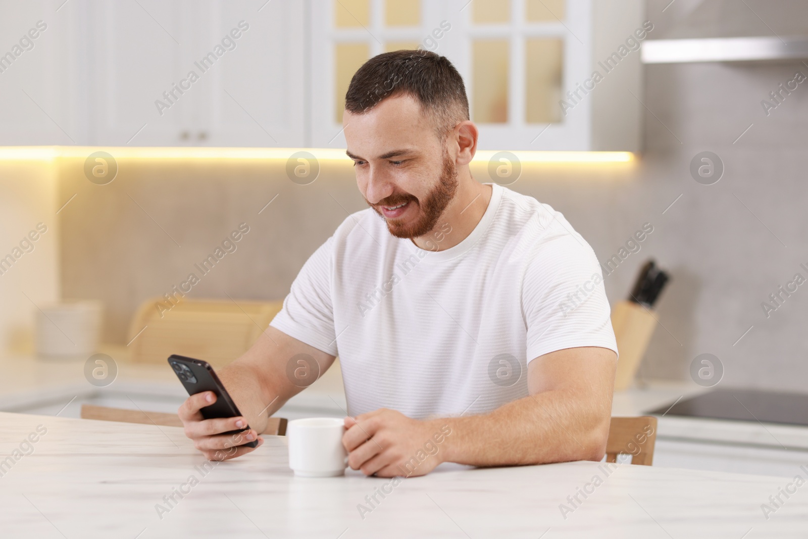 Photo of Happy man using smartphone at white table in kitchen