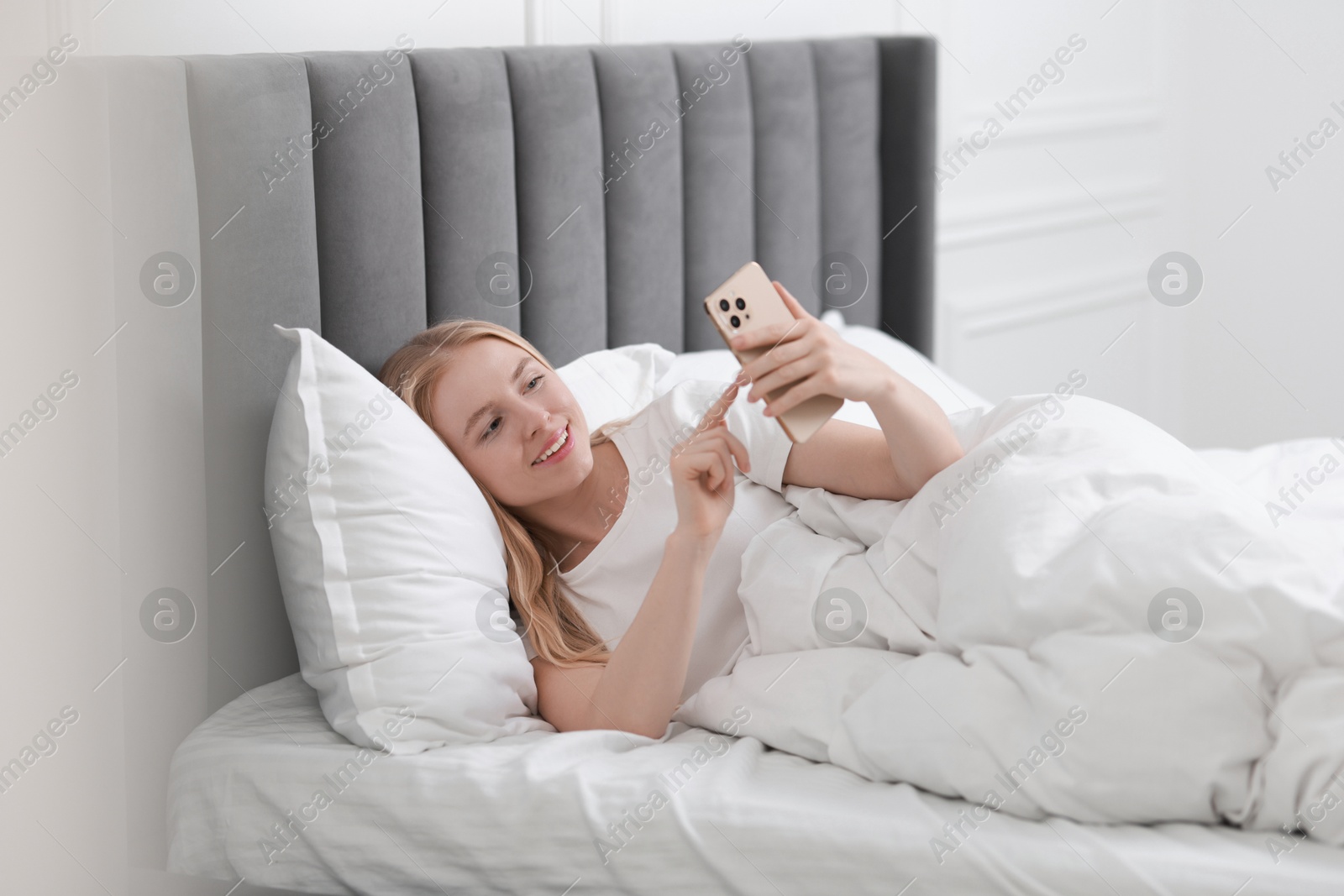 Photo of Smiling woman looking at smartphone in bed