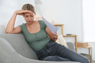 Photo of Menopause. Woman waving paper sheet to cool herself during hot flash on sofa at home