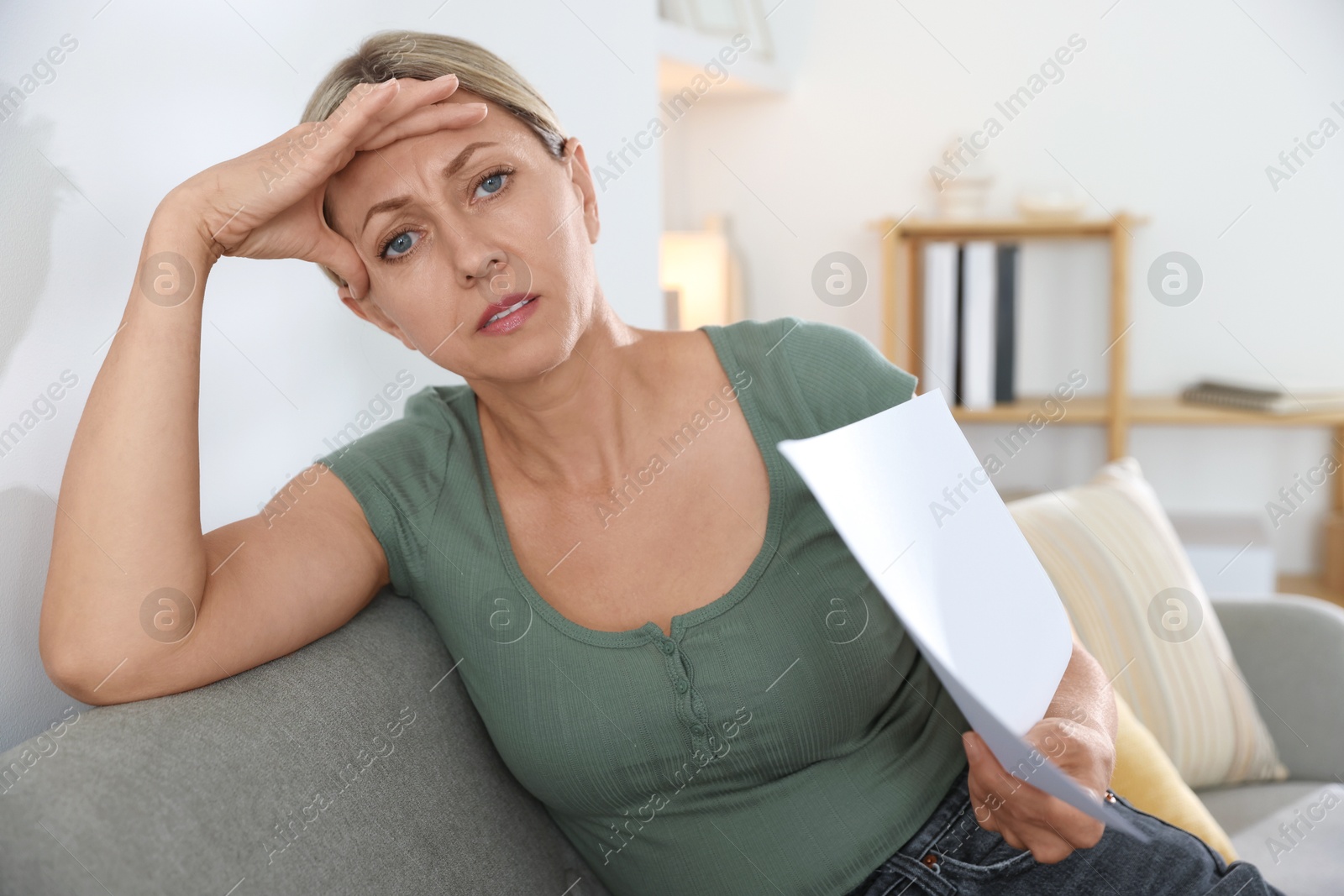 Photo of Menopause. Woman waving paper sheet to cool herself during hot flash on sofa at home