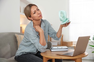 Photo of Menopause. Woman using electric fan to cool herself during hot flash at table indoors