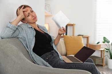 Photo of Menopause. Woman waving paper sheet to cool herself during hot flash on sofa at home