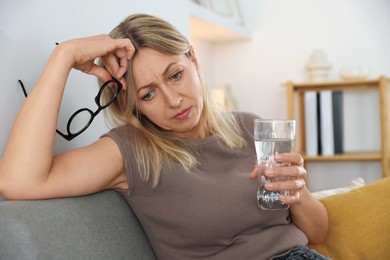 Photo of Menopause. Woman with glass of water suffering from headache on sofa at home