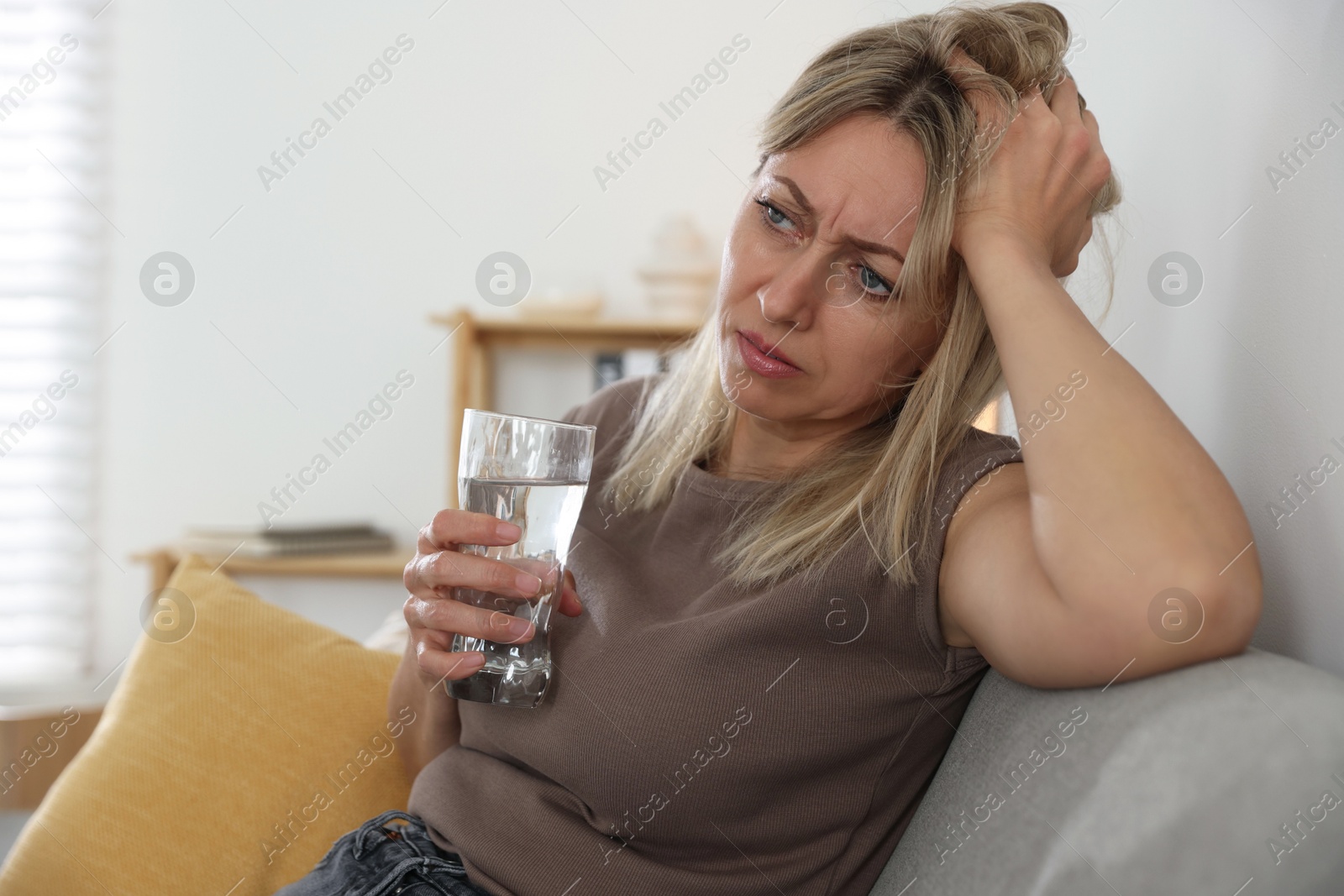 Photo of Menopause. Woman with glass of water suffering from headache on sofa at home