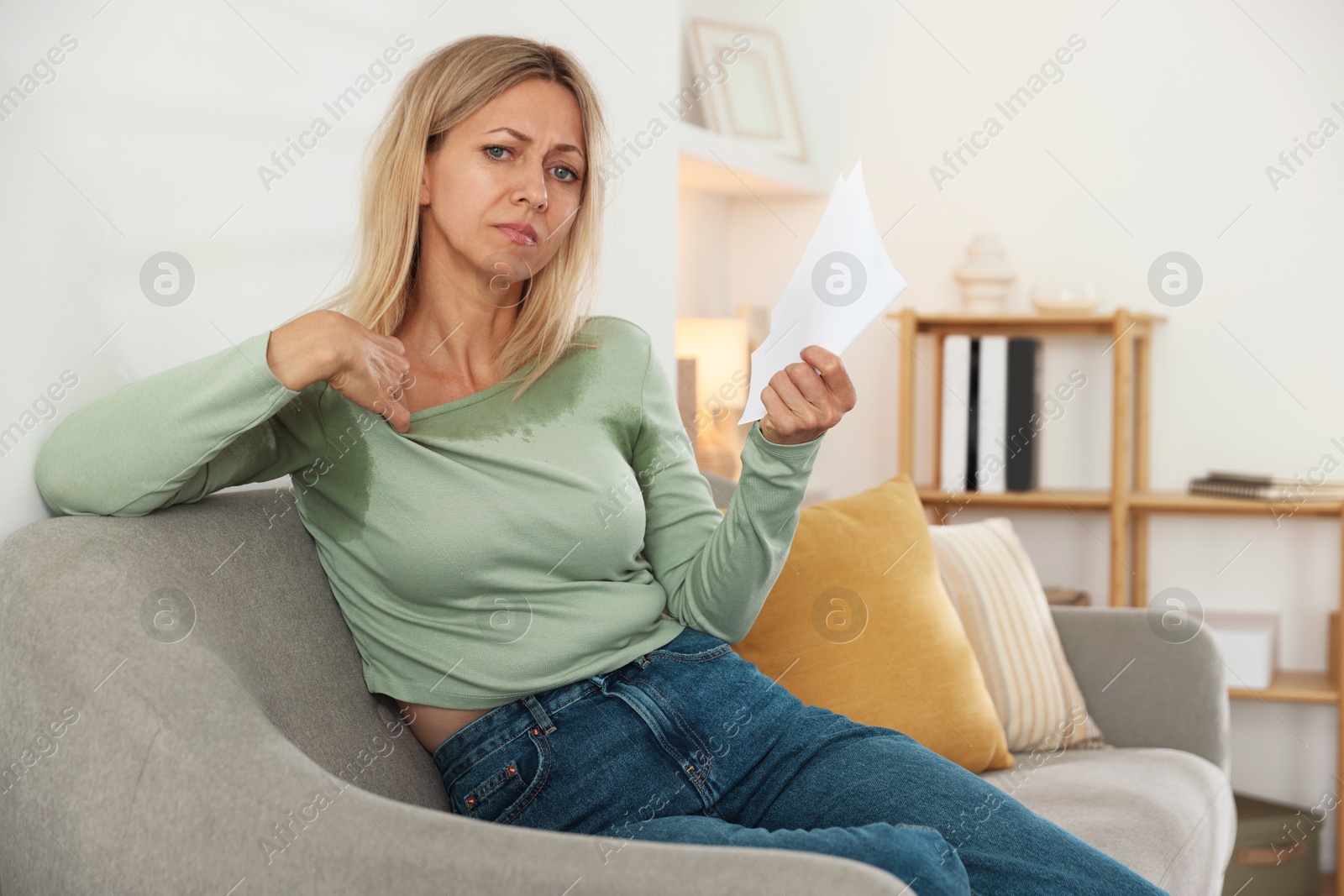 Photo of Menopause. Woman waving paper sheet to cool herself during hot flash on sofa at home