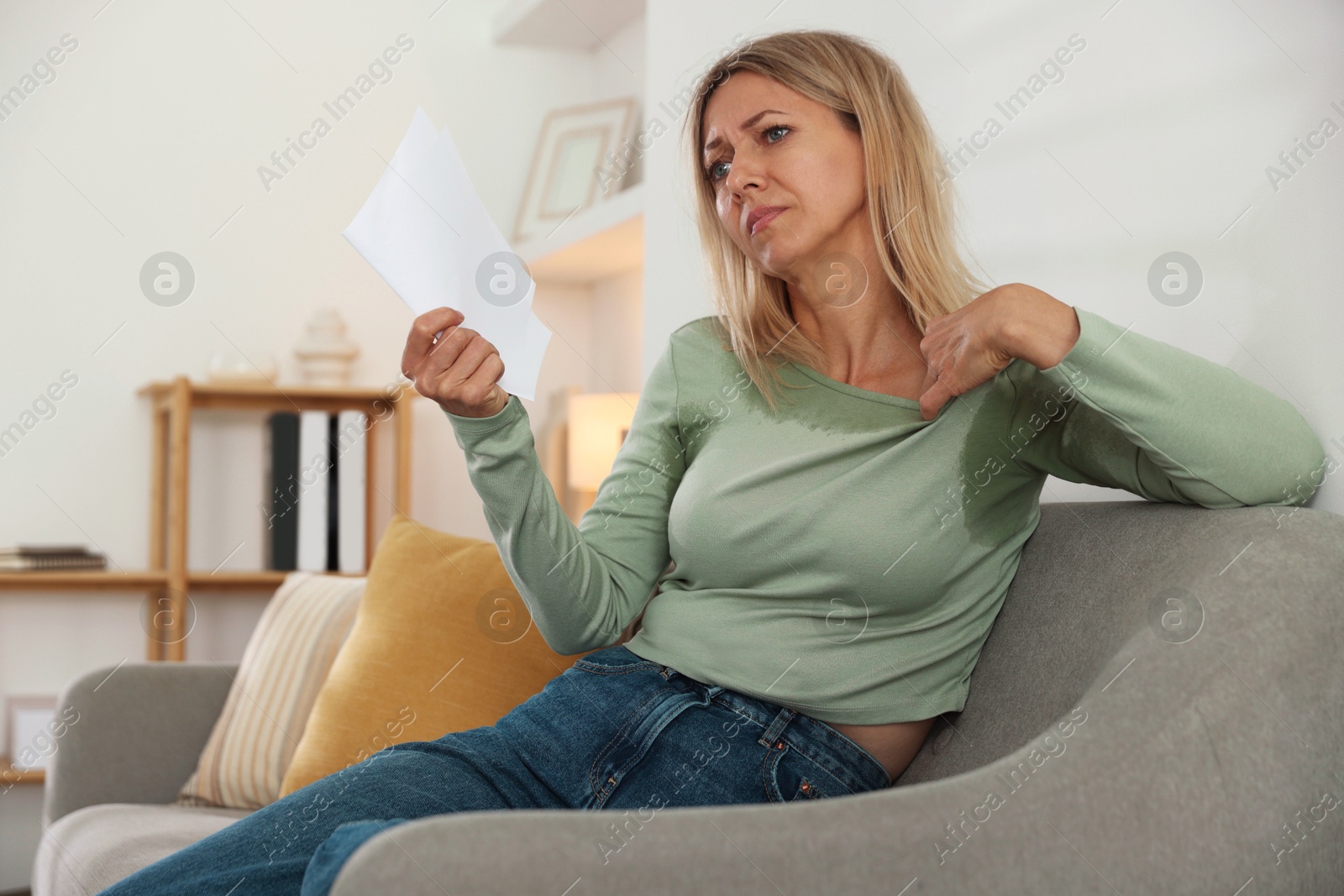 Photo of Menopause. Woman waving paper sheet to cool herself during hot flash on sofa at home